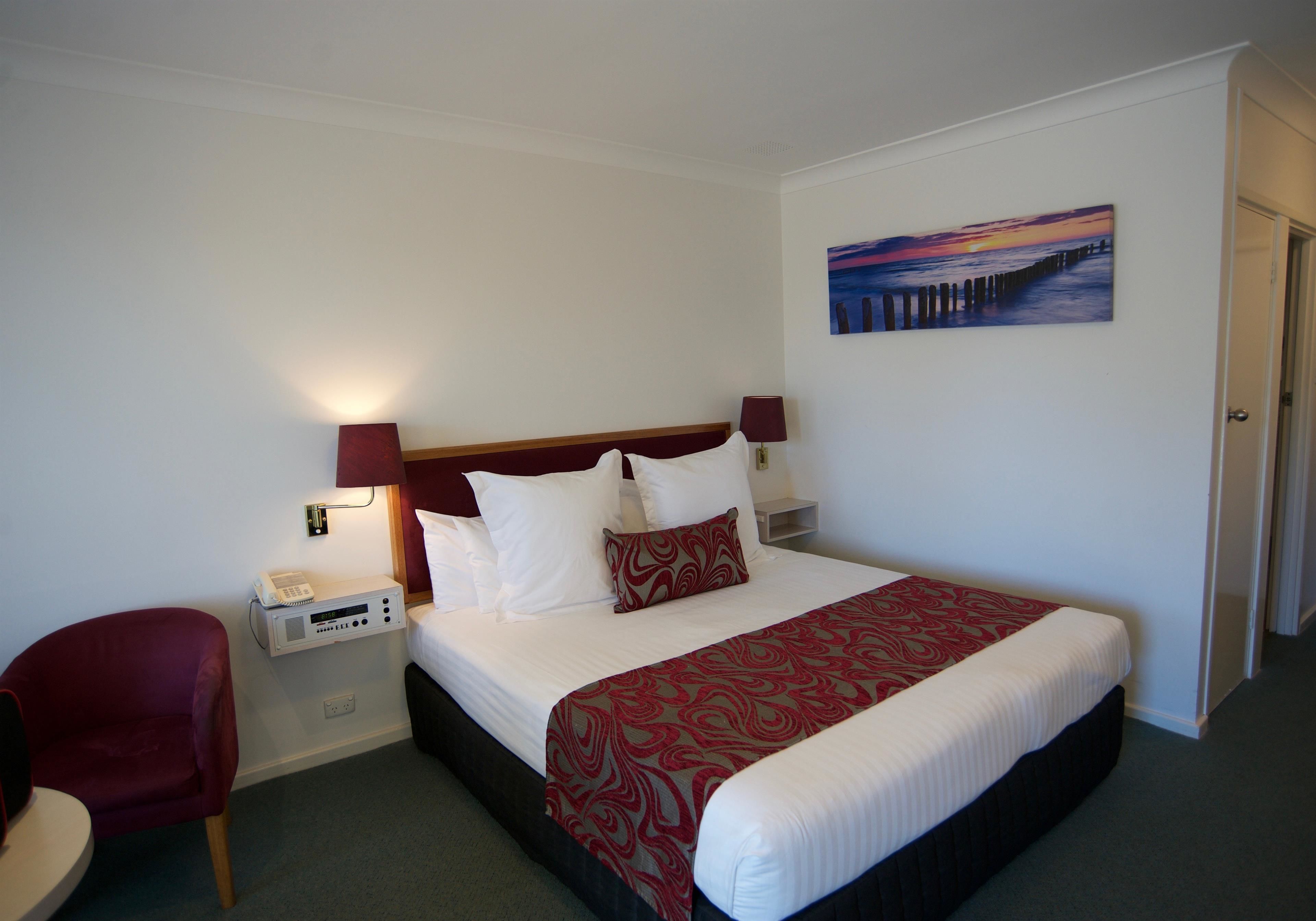 A hotel room with a bed, a red chair, a bedside table with a phone, and a painting on the wall.