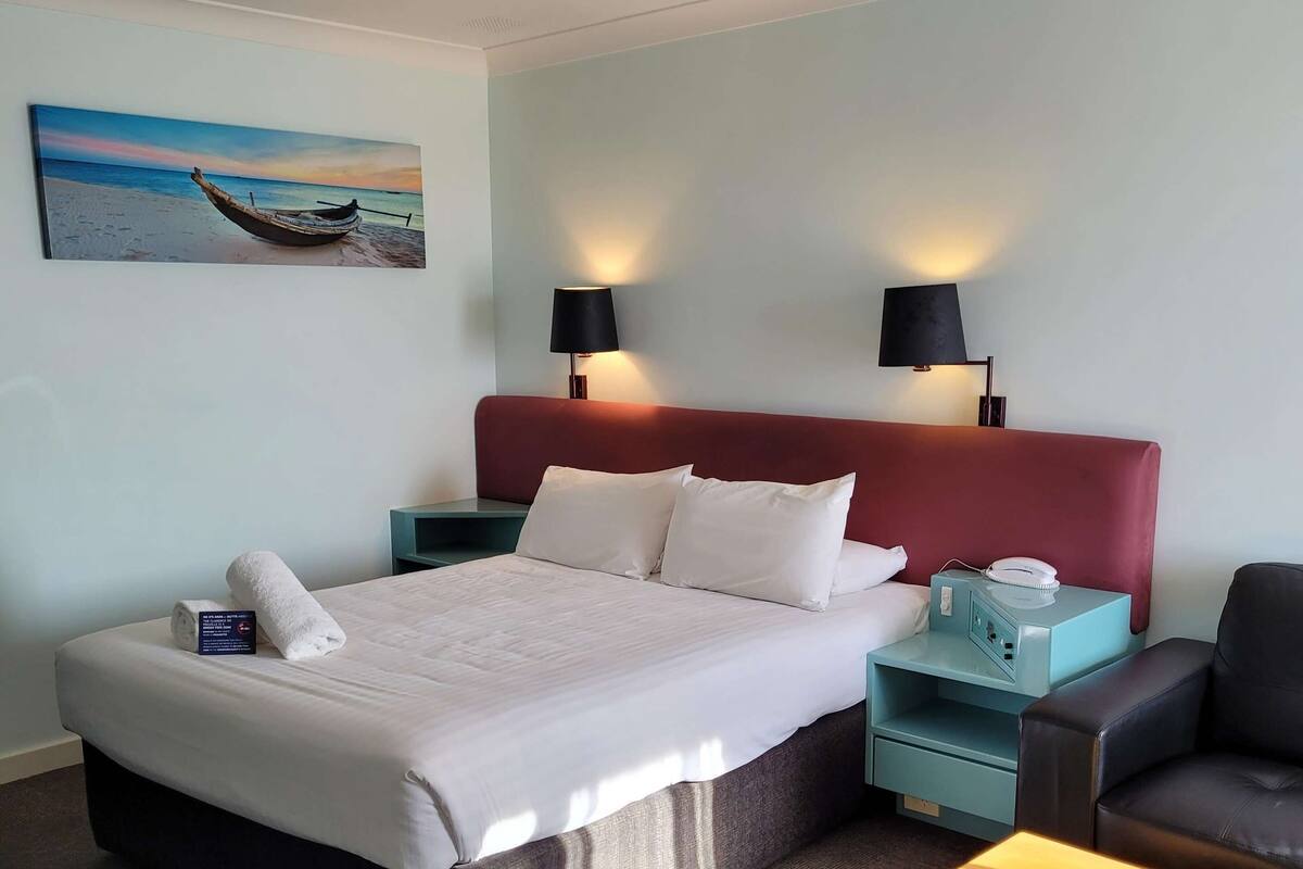 Ein Hotelzimmer mit einem Bett, zwei Nachttischen, einem Sessel und einem kleinen Tisch, sowie einem Gemälde an der Wand.