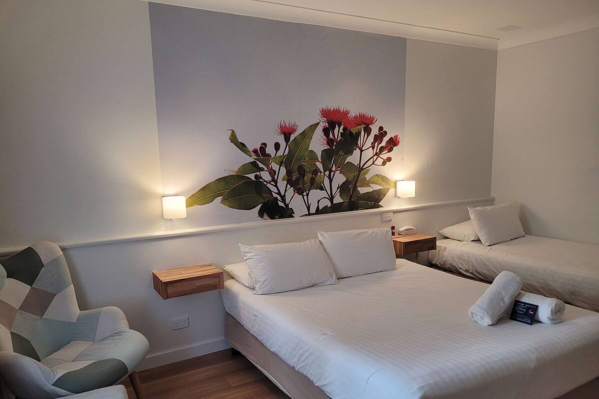 Ein Hotelzimmer mit zwei Betten, einer Schlafcouch, einer Wandpflanze und Nachttischlampen.