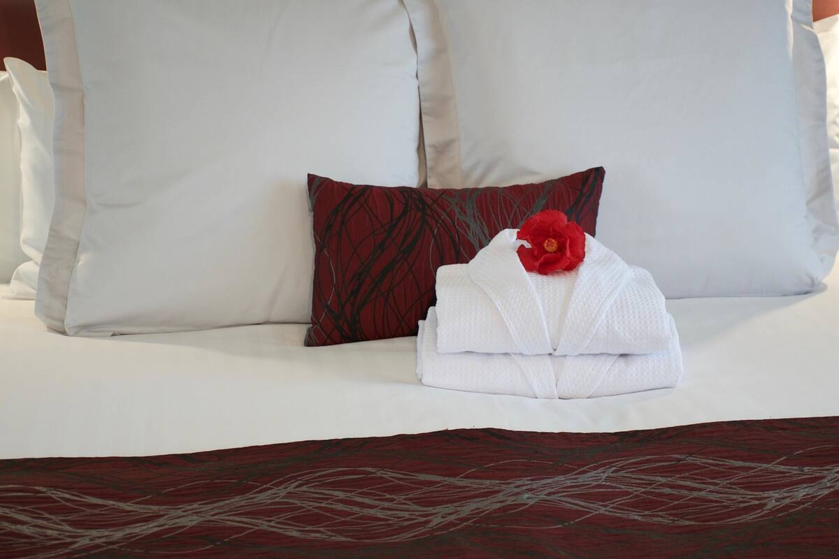 Ein ordentlich bezogenes Bett mit weißen Leinen, einem Kissen in Rot-Schwarz-Muster und einem gefalteten Handtuch mit einer roten Blume.