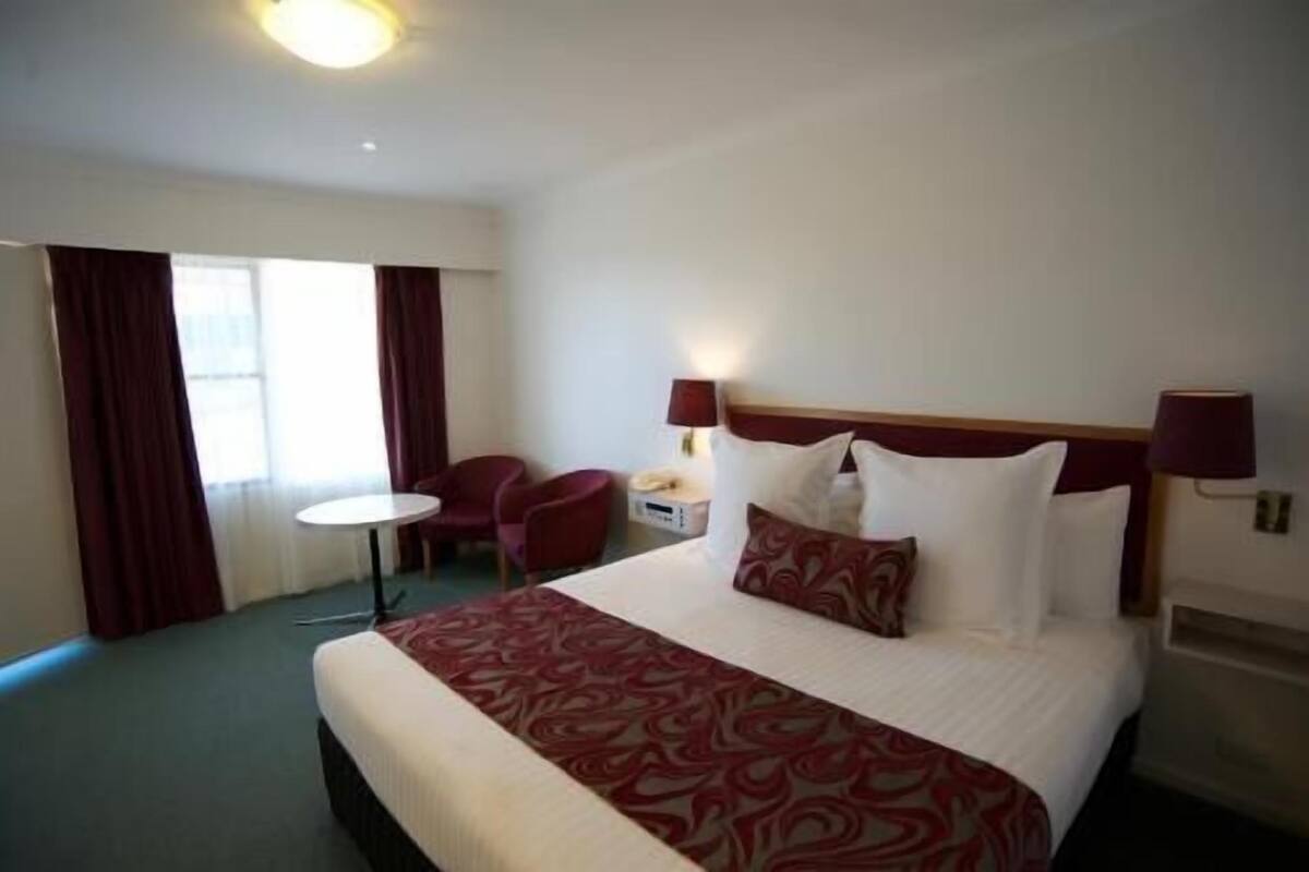A hotel room with a bed, two bedside lamps, a small table, a chair, and a window with curtains.