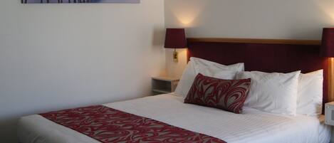 A hotel room with a large bed, a red and white patterned bedspread, two bedside lamps, and a painting of a pier on the wall.
