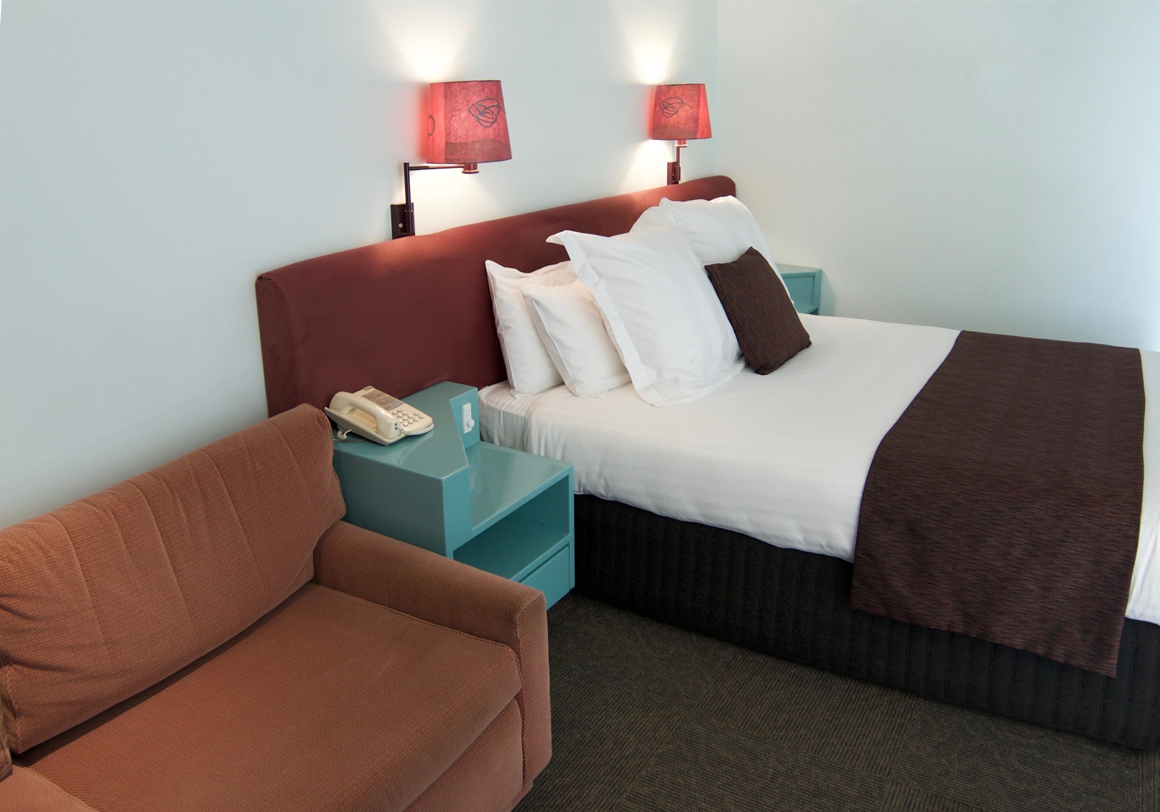 A hotel room with a bed, a red headboard, two bedside lamps, a telephone, a teal nightstand, and a brown sofa.
