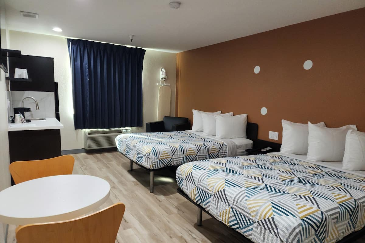 A hotel room with two beds, a small table, a chair, a desk with a sink, and a window with curtains.