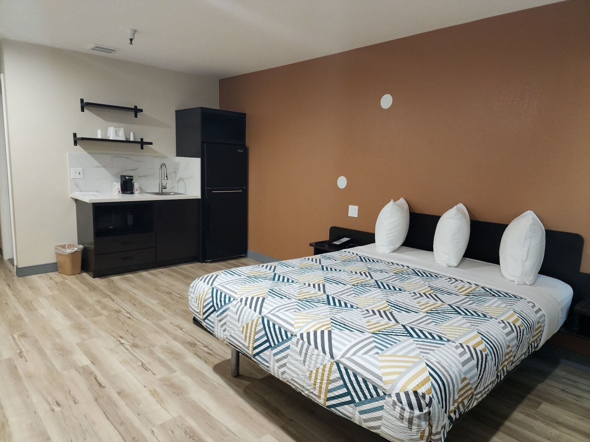 A bedroom with a bed, a nightstand, a dresser, a built-in desk with a chair, and a kitchenette.
