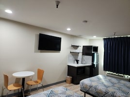 Habitación compacta con kitchenette, televisor de pantalla plana en la pared, una pequeña mesa redonda con dos sillas y una cama con ropa de cama estampada.