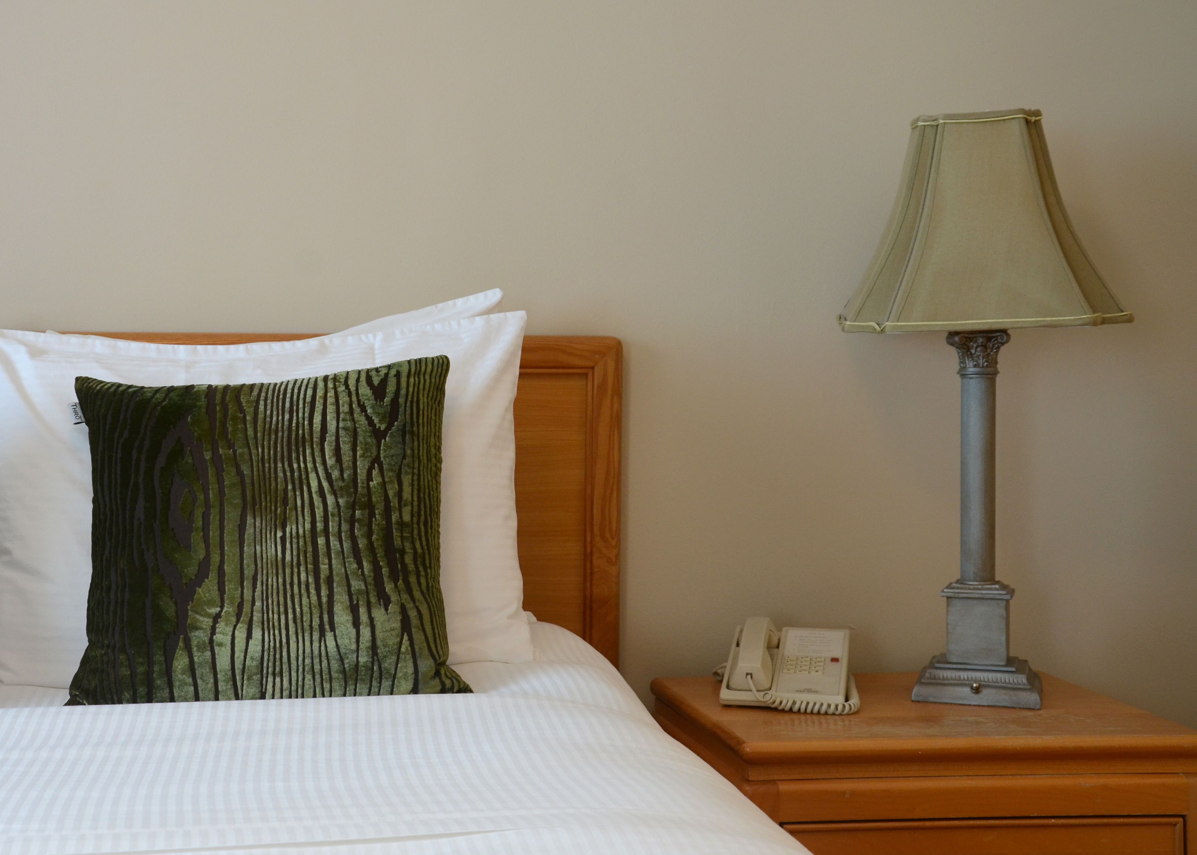 A neatly made bed with white linens and a green patterned pillow, a bedside lamp, and a telephone on a wooden nightstand.