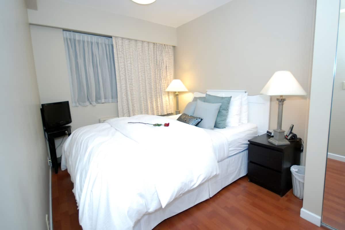 Una cama bien hecha con sábanas blancas, una mesita de noche con lámpara, un televisor sobre un mueble bajo y una ventana con cortinas traslúcidas.