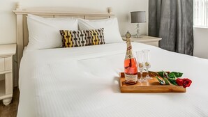 Un lit bien fait, avec du linge de lit blanc, une table de chevet en bois surmontée d’une lampe, et une autre table de chevet avec une bouteille de champagne Henkell, des verres et une rose.