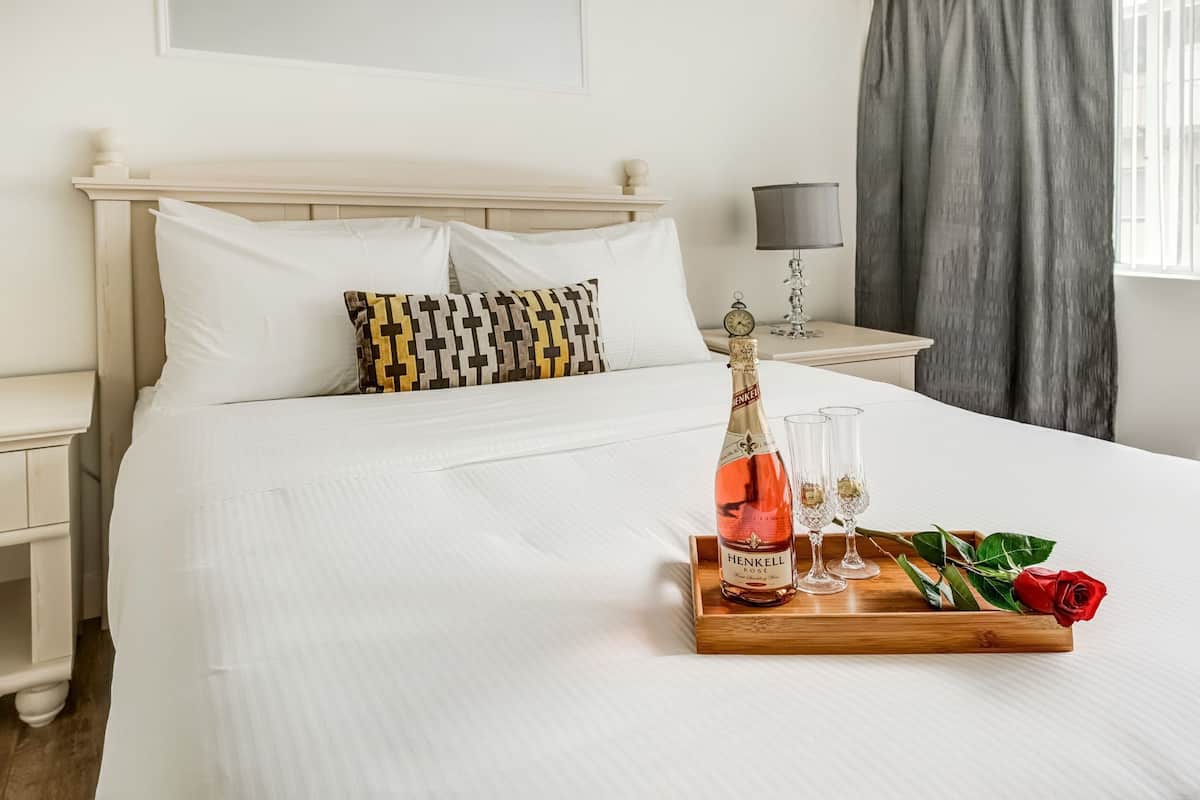 Una cama bien hecha con sábanas blancas, una mesita de noche de madera con una lámpara, y otra mesita de noche con champán Henkell, copas y una rosa.