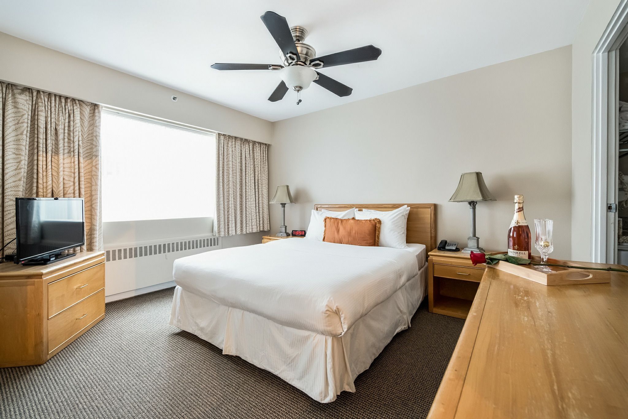 A hotel room with a large bed, a ceiling fan, a TV on a wooden dresser, and a window with curtains.