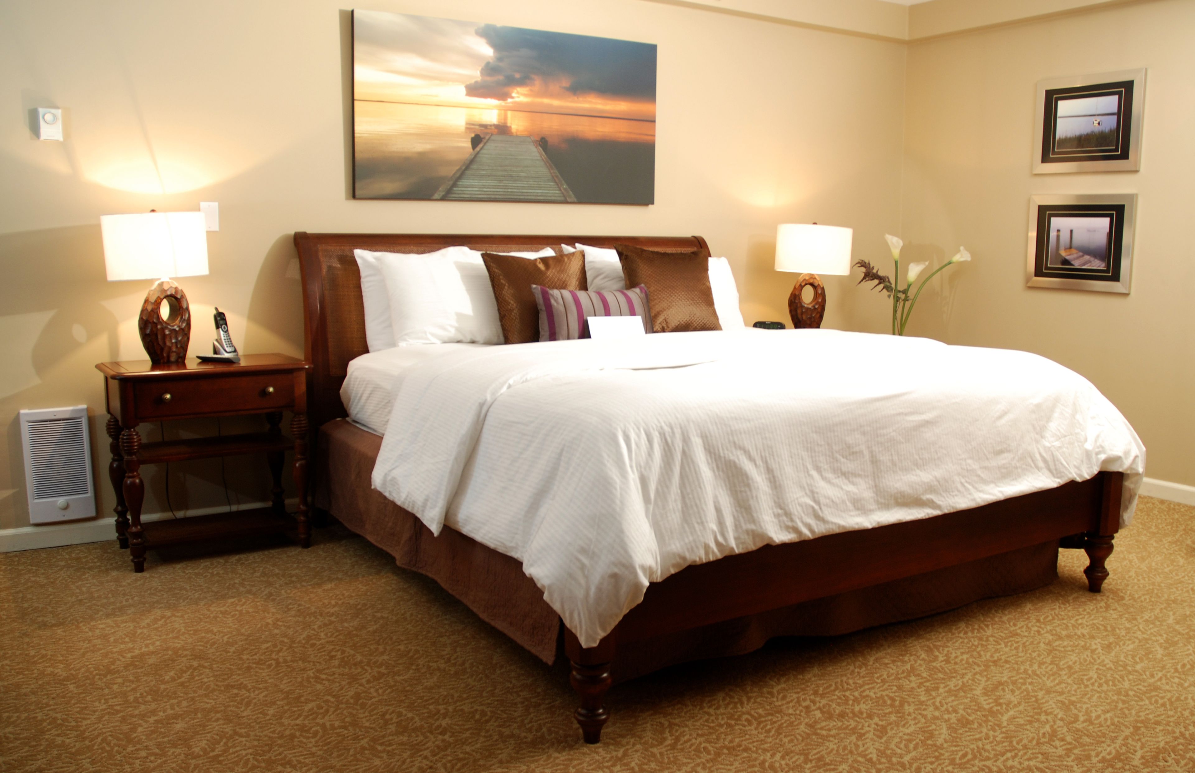 A hotel room with a large bed, two bedside tables, a nightstand, a wall-mounted lamp, a painting, and a framed picture.