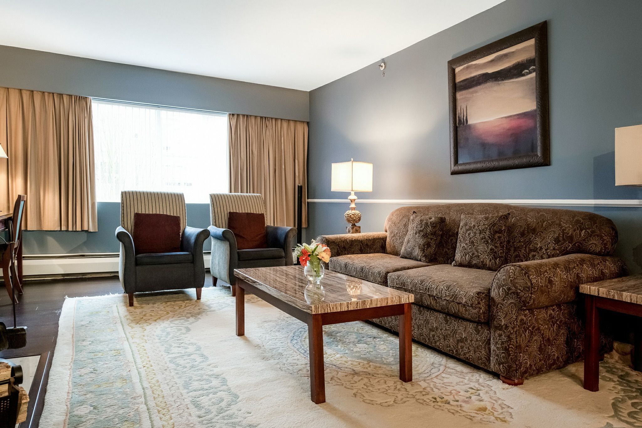 A living room with a patterned sofa, a coffee table, and a chair. There is a lamp and a painting on the wall.