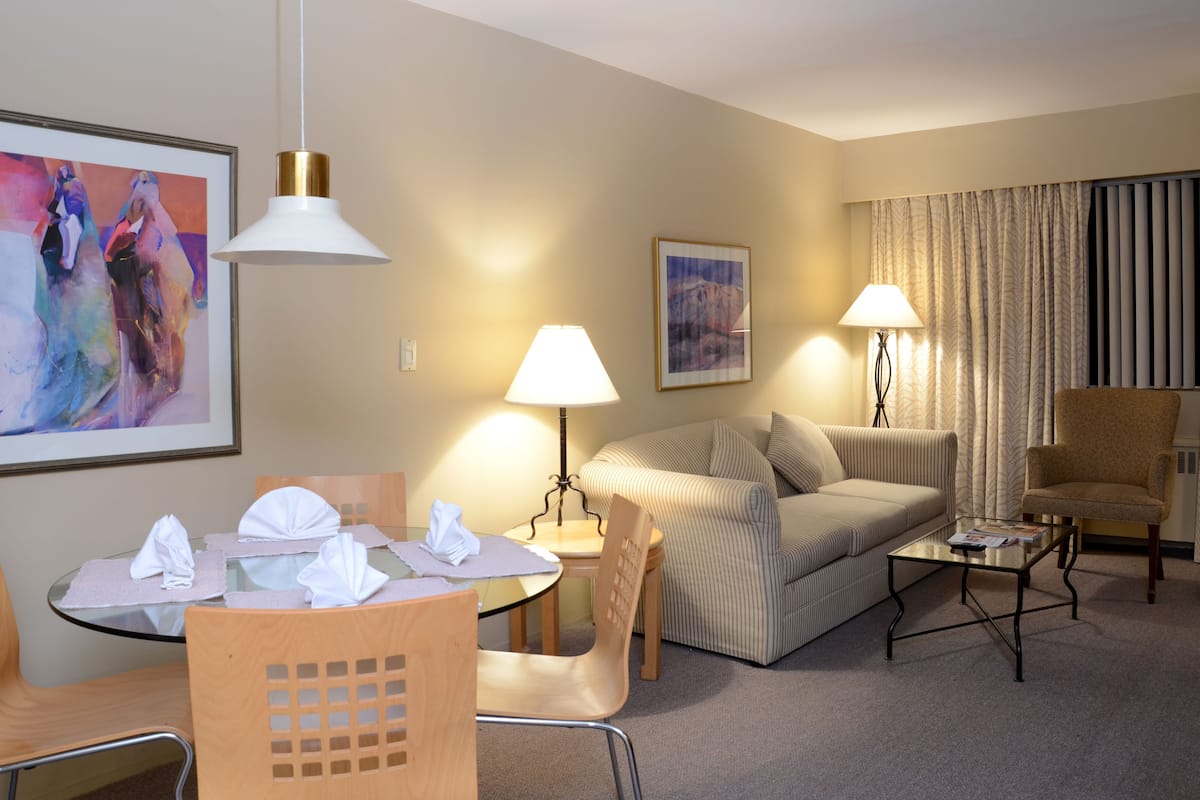 Habitación de hotel con zona de comedor, sofá, silla, lámpara y un cuadro en la pared.