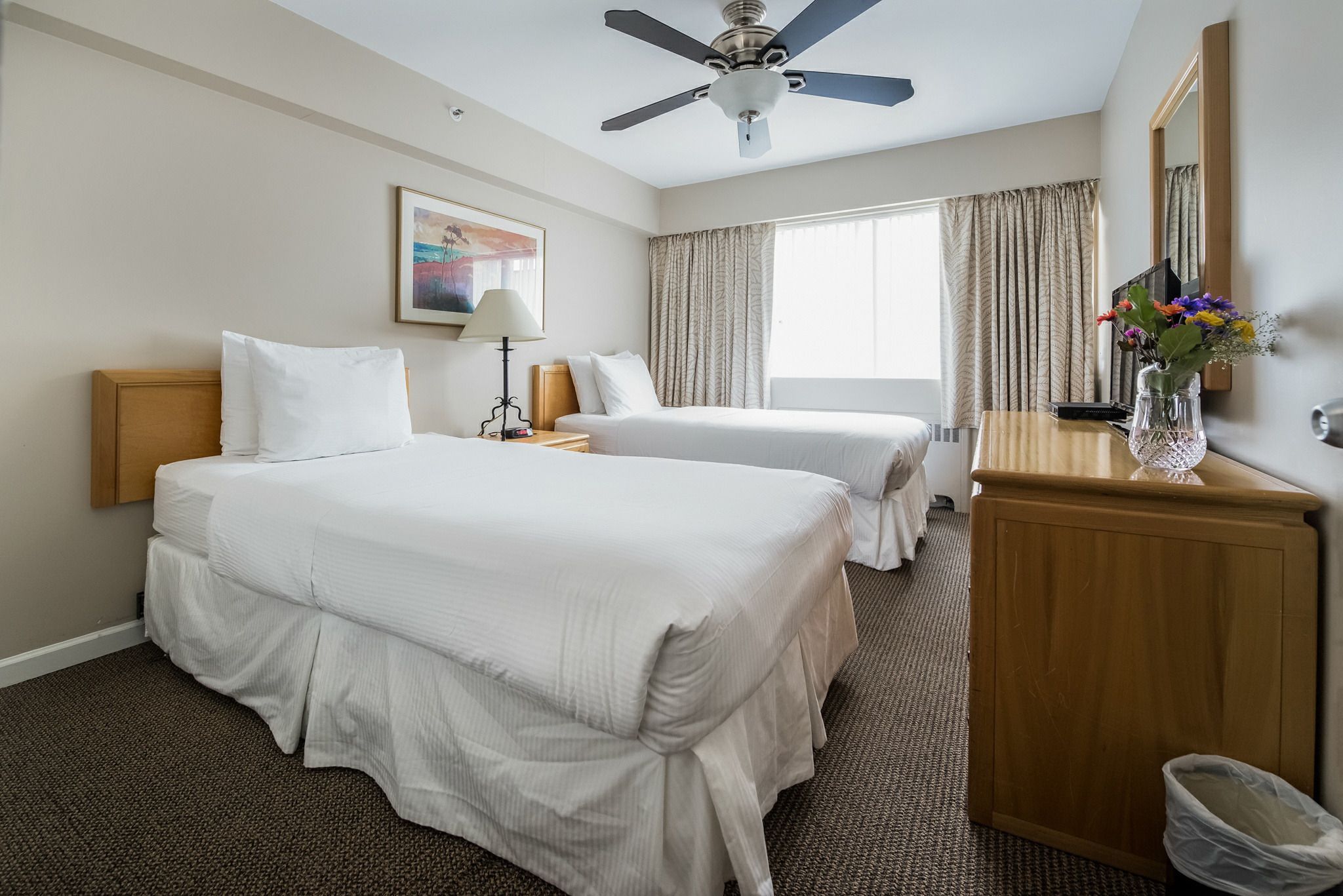 A hotel room with two beds, a ceiling fan, a large window with curtains, a wooden cabinet, and a framed picture on the wall.