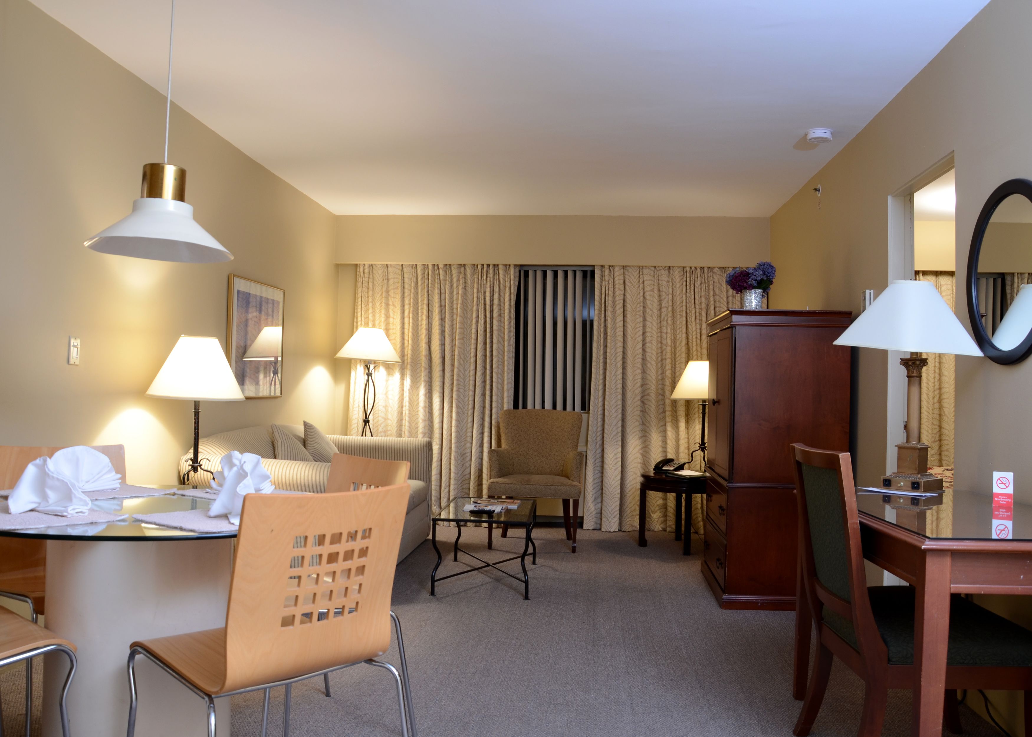 A hotel room with a dining table, chairs, a sofa, a chair, a desk, lamps, a mirror, and a cabinet.