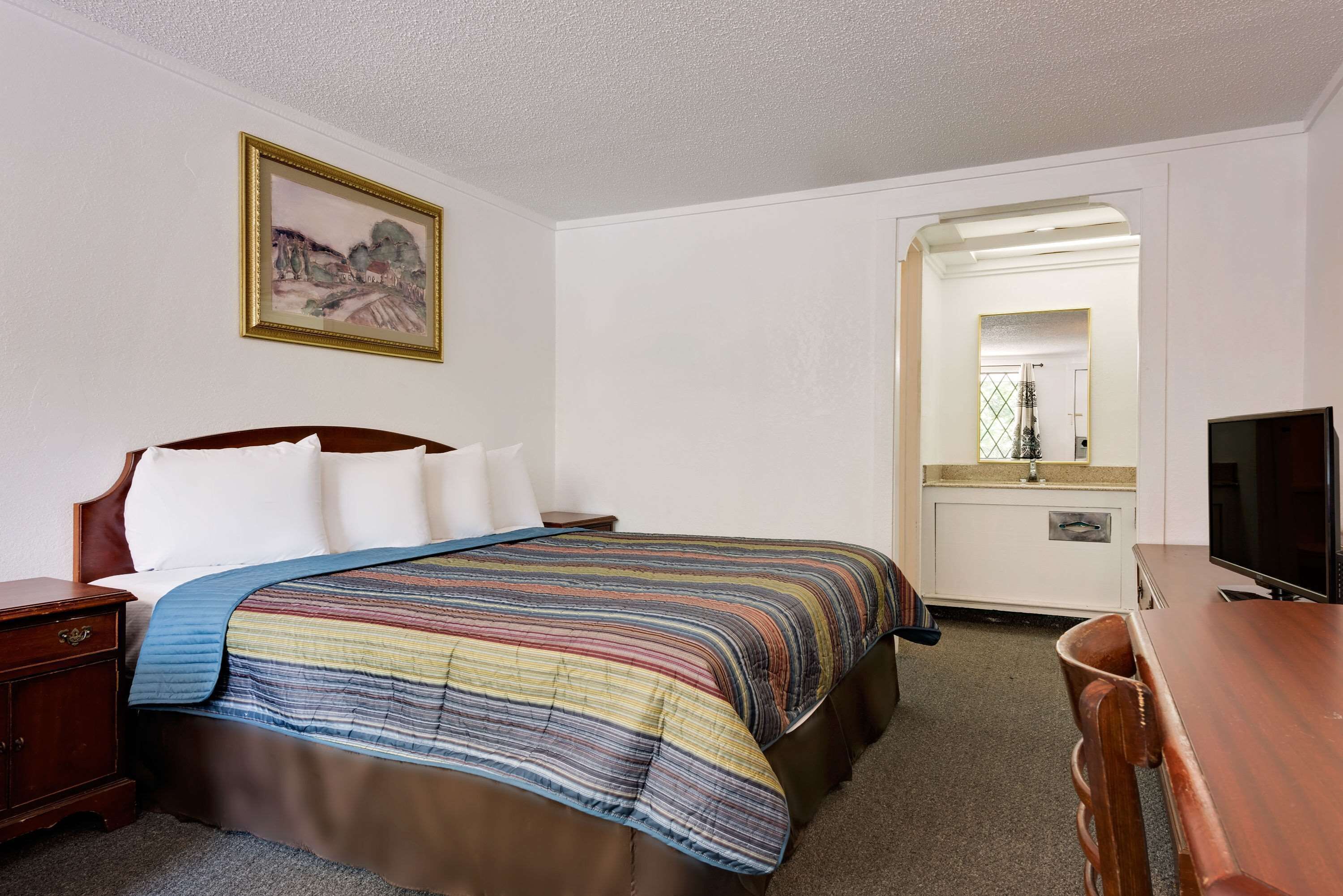 A hotel room with a bed, a wooden nightstand, a desk, a television, and a mirror.