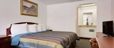 Una habitación de hotel con una cama, una mesita de noche de madera, un escritorio, un televisor y un espejo.