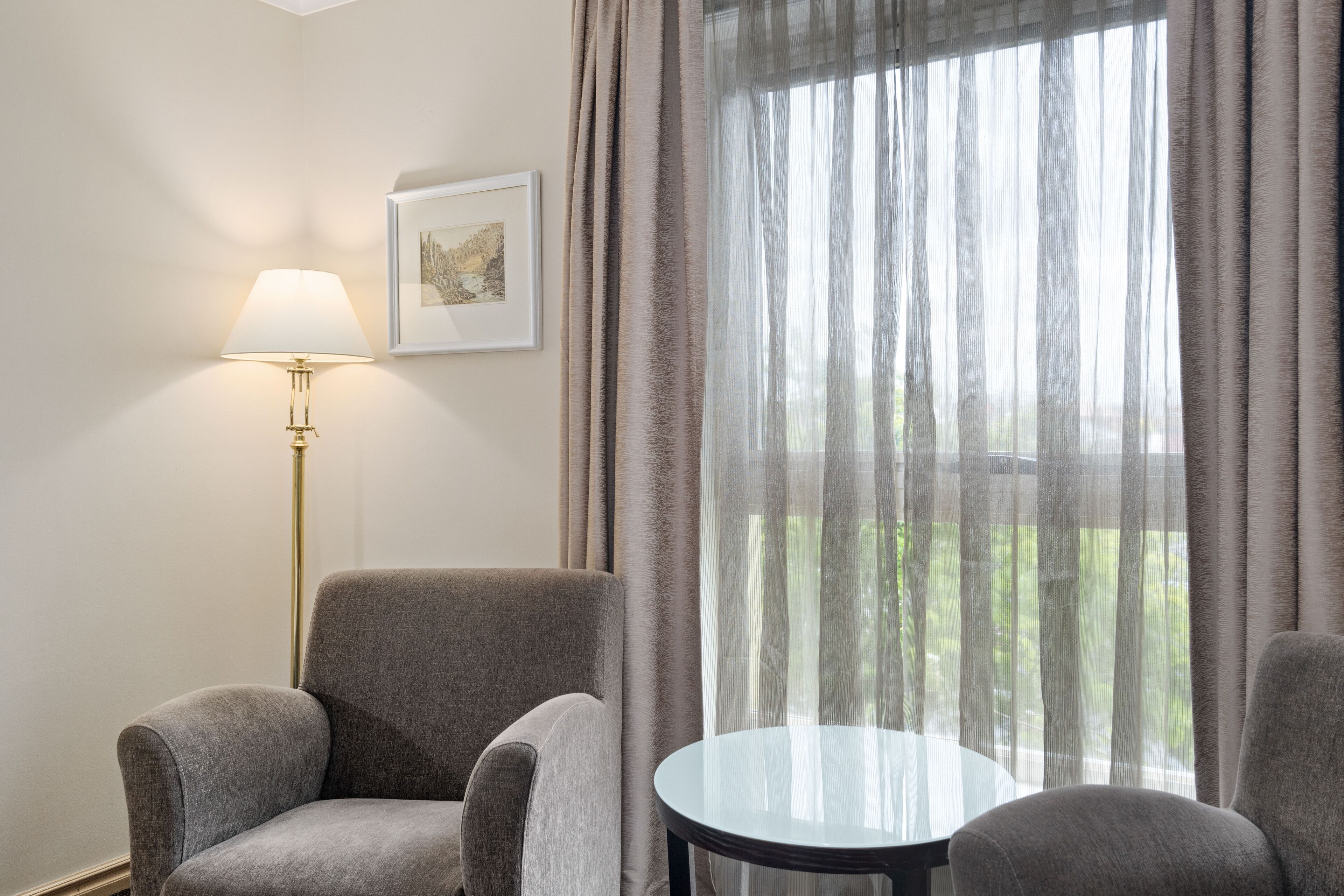 Una habitación con dos sillones grises, una mesita redonda pequeña, una lámpara de pie y una ventana con cortinas traslúcidas.