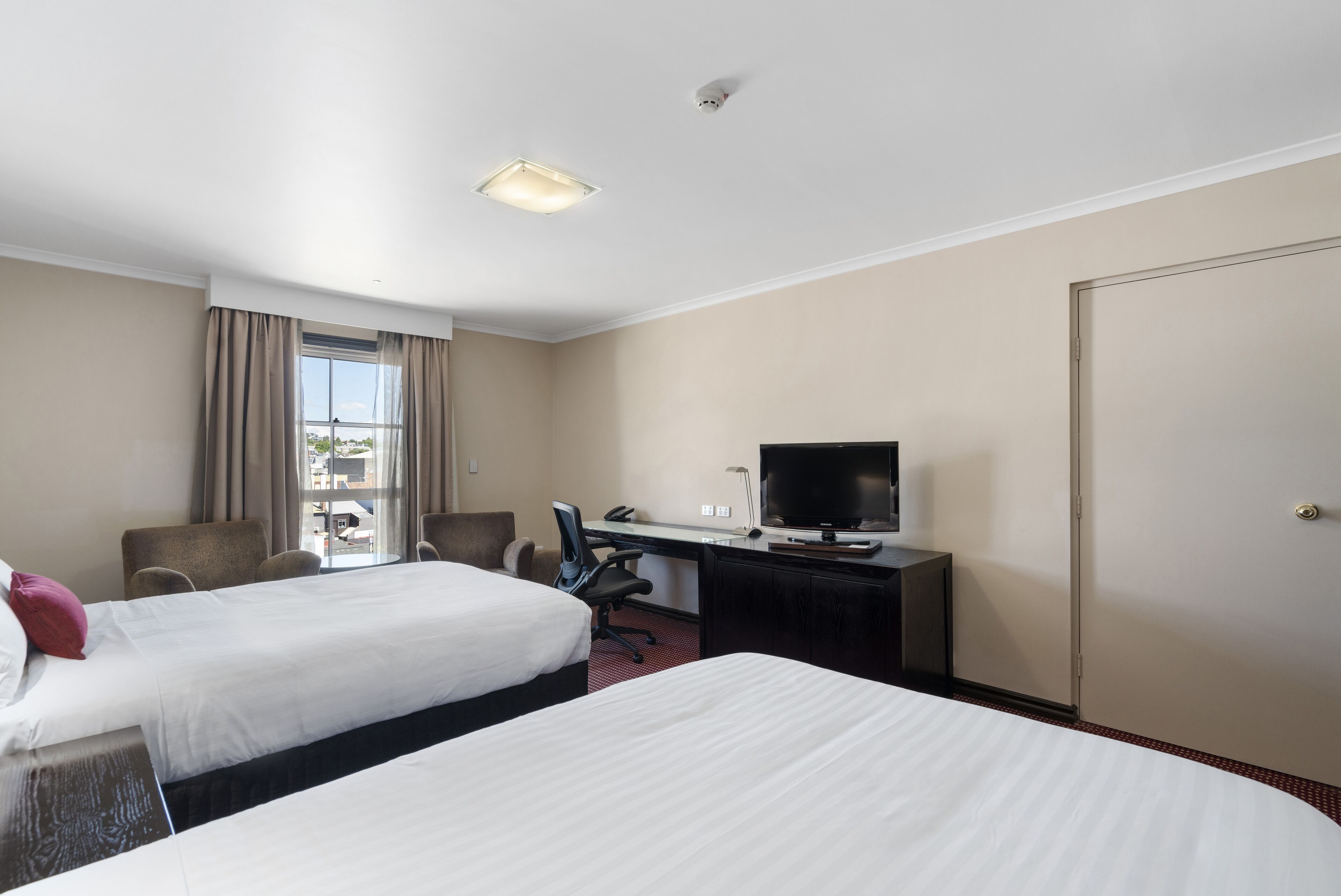 Quarto de hotel com duas camas, uma secretária com televisão, uma cadeira e uma janela com cortinas.
