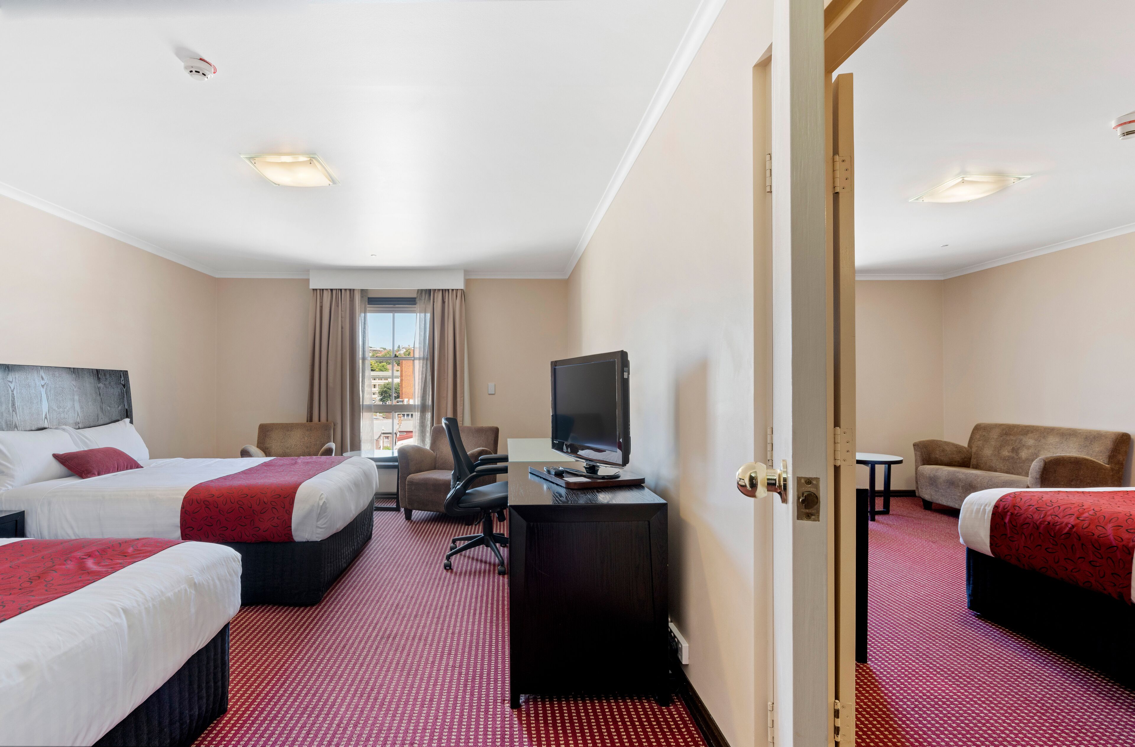 Habitación de hotel con tres camas, un escritorio con televisor, una silla y una mesita.