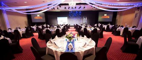 Un salón de banquetes con mesas redondas dispuestas para un evento formal, con un escenario que incluye una pantalla mostrando el logotipo del hotel.
