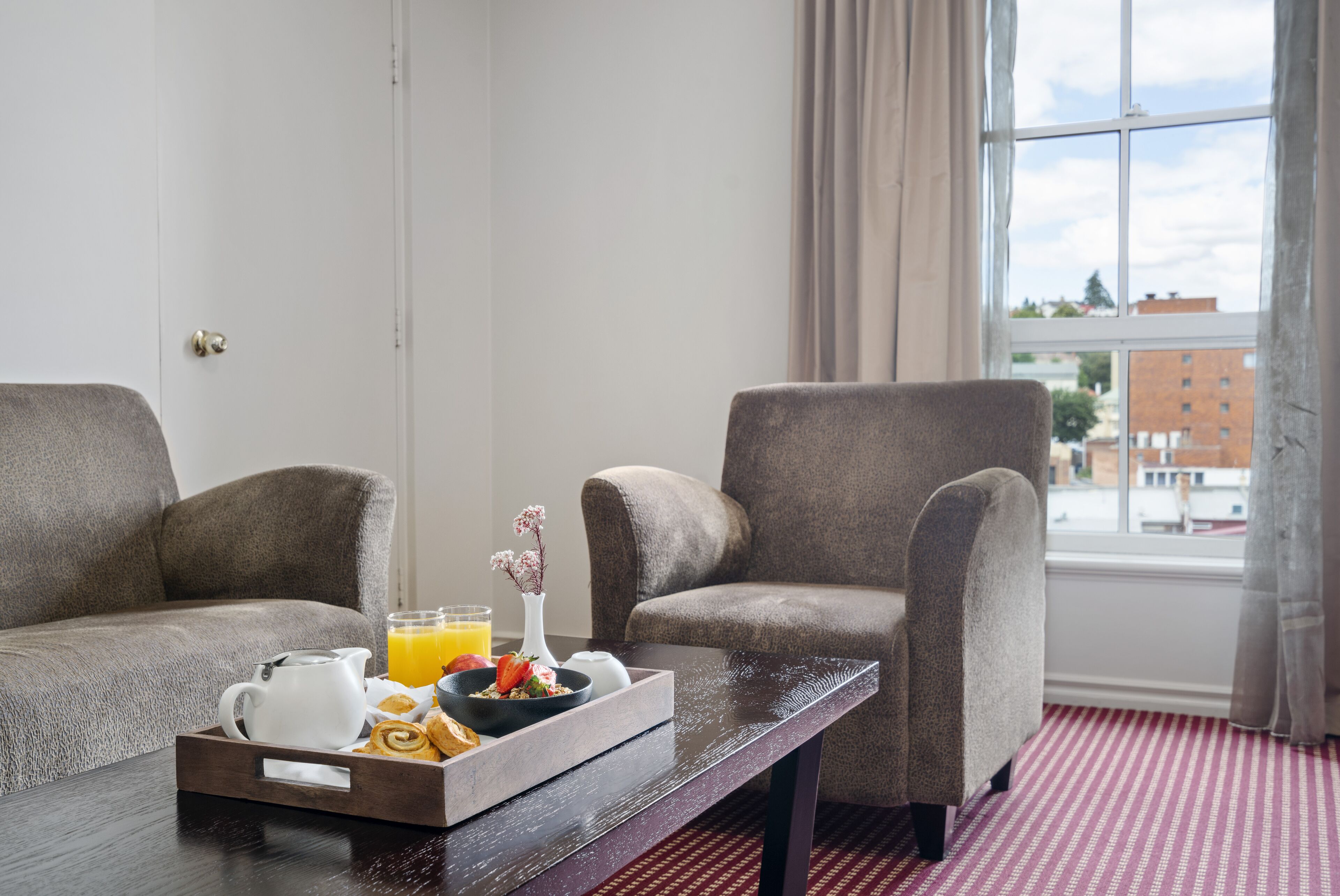 A room with a sofa, armchair, breakfast tray, and a window with a view of buildings.