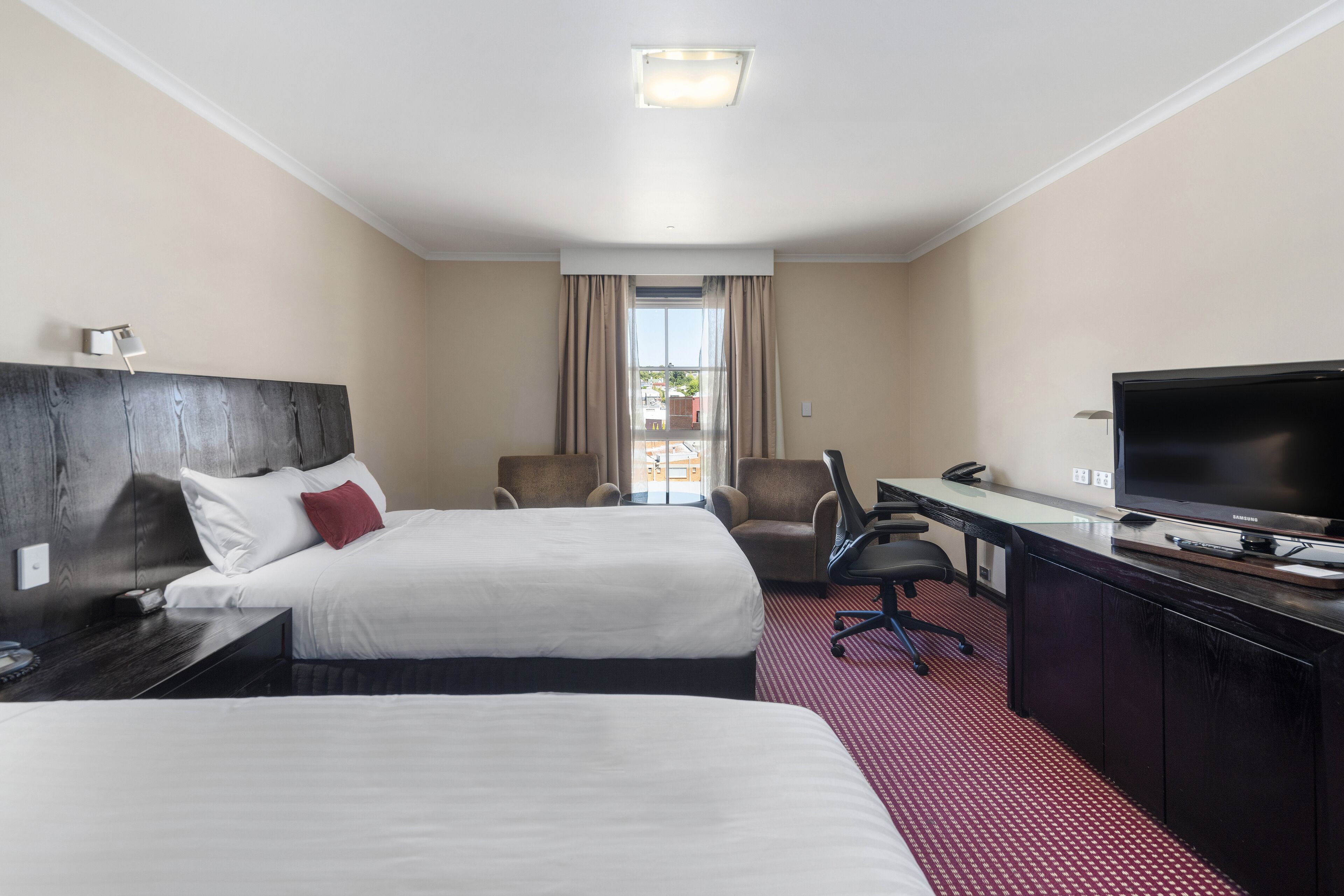 Habitación de hotel con dos camas, un escritorio con televisor, una silla y vistas al exterior.
