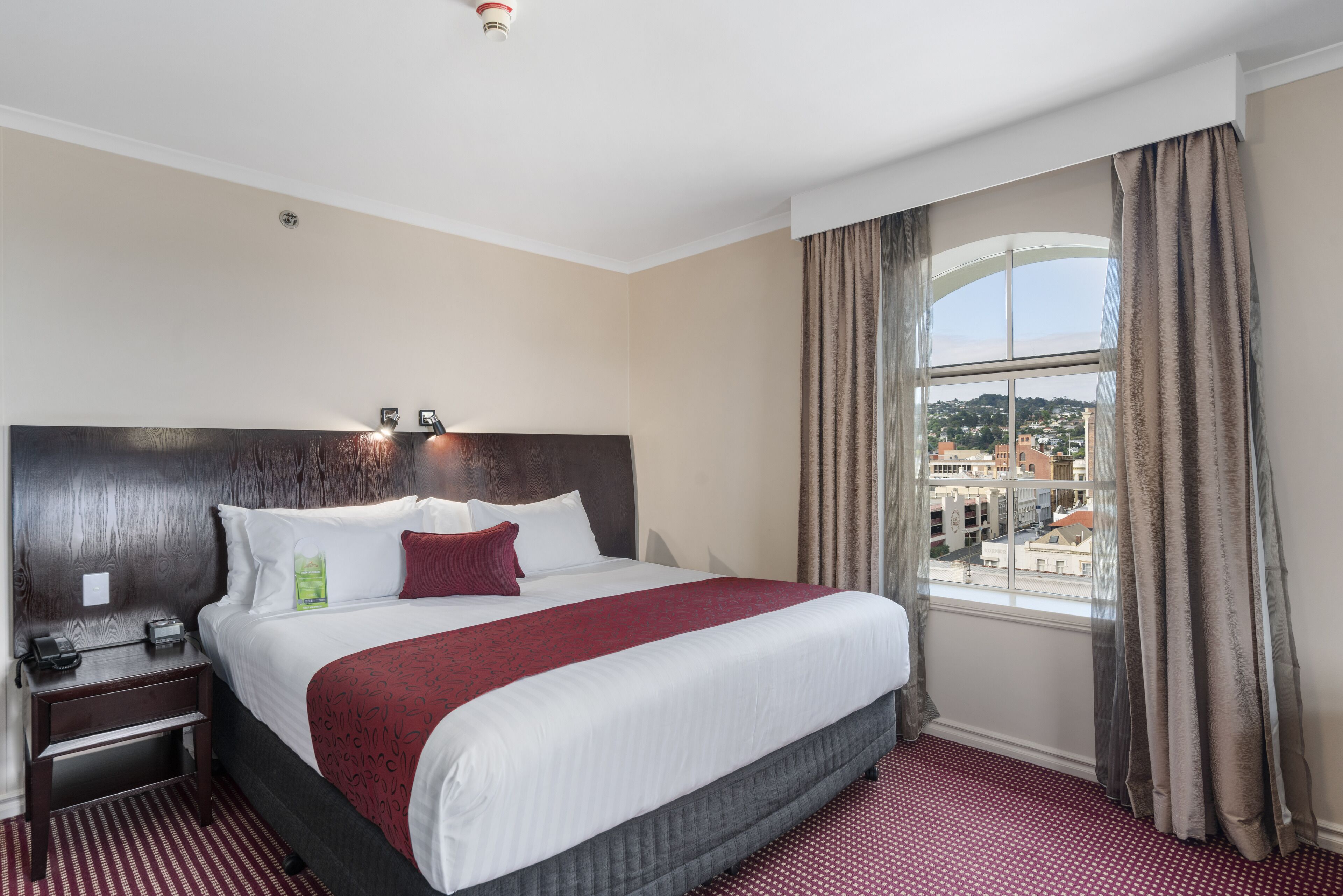Habitación de hotel con una cama grande, una mesita de noche, un teléfono y una ventana con vistas a edificios.