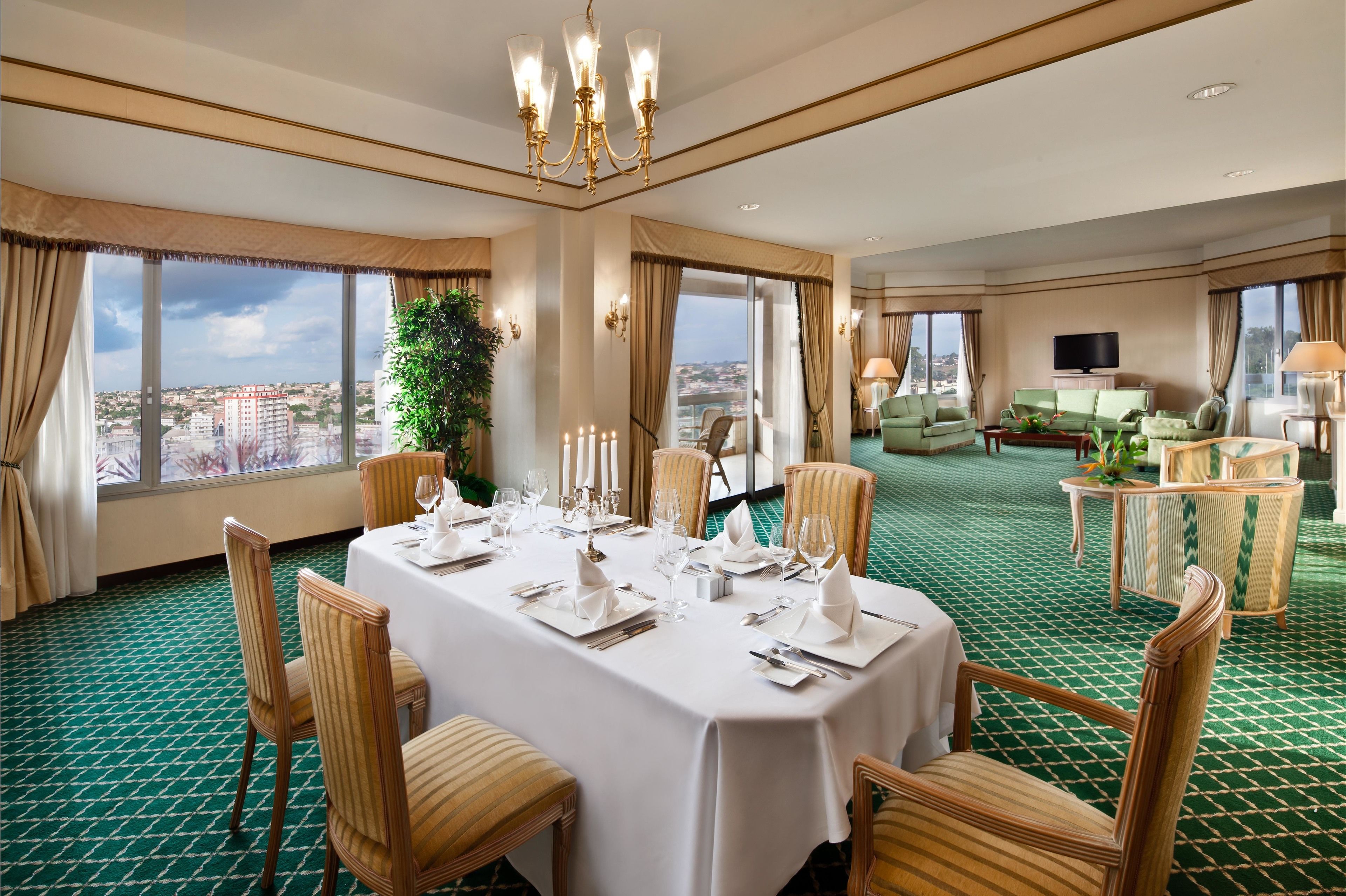 Una camera d'albergo con zona pranzo apparecchiata per un pasto, ampie finestre con vista sulla città e un'area living con televisione e posti a sedere.