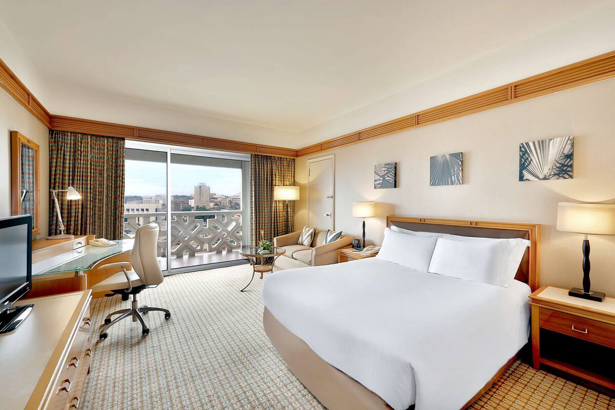A hotel room with a large bed, a desk with a computer, a chair, a sofa, and a balcony with a view of the city.