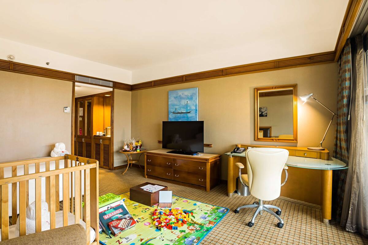 A hotel room with a crib, a desk with a computer, a television, and a large window with curtains.
