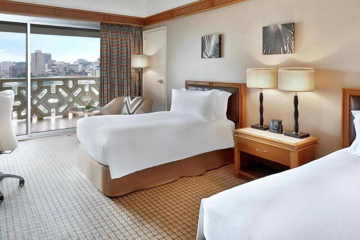 A hotel room with a bed, a chair, a nightstand with lamps, and a view of the city through a balcony.