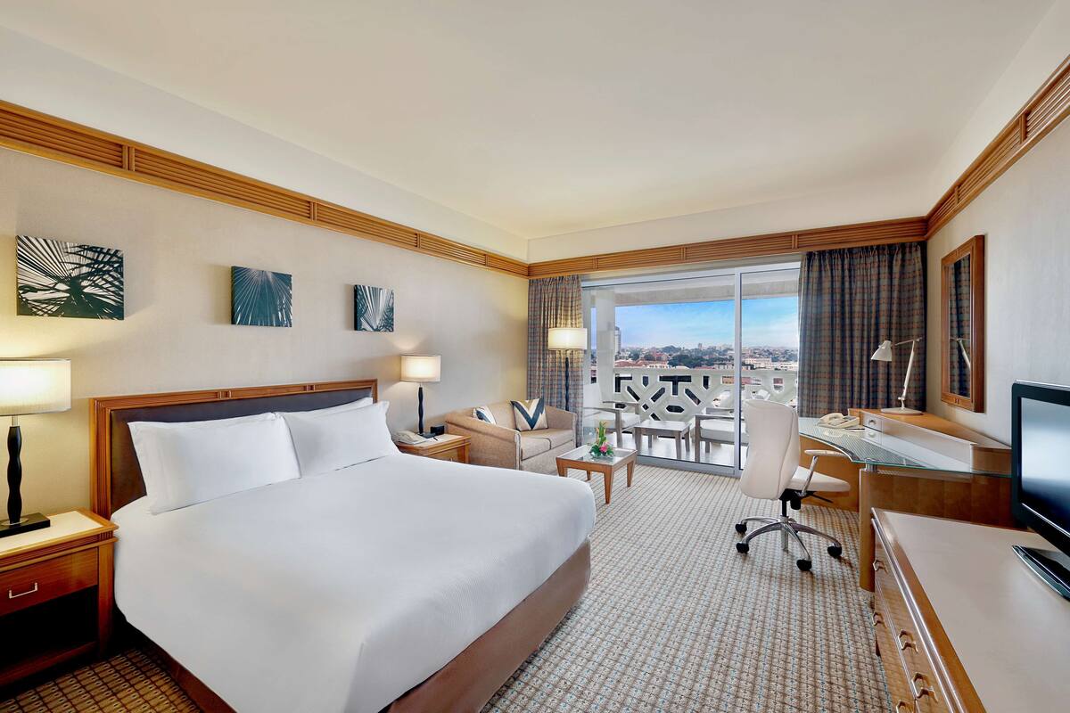 A hotel room with a large bed, a desk with a computer, a chair, a desk lamp, and a view of the city through the window.