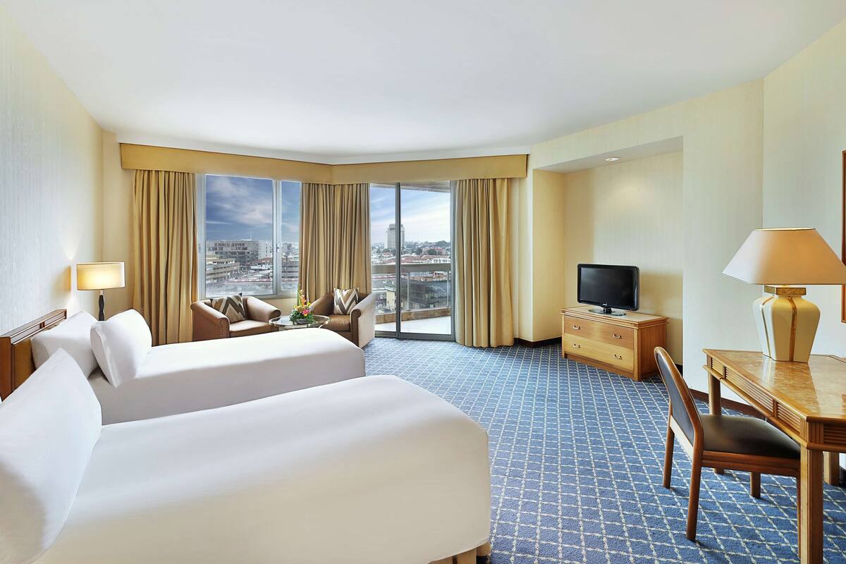 A hotel room with two beds, a desk with a TV, and a city view through large windows.