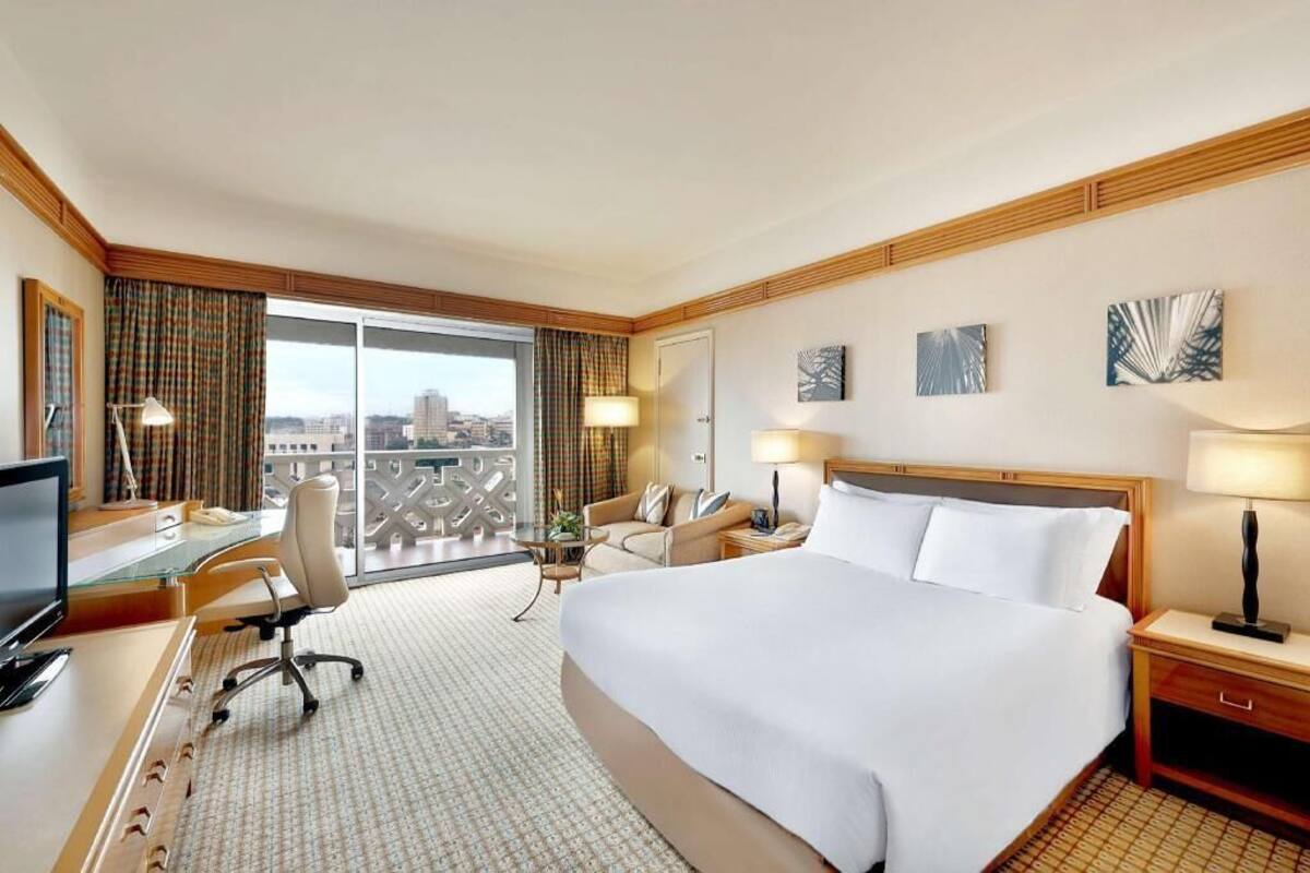 A hotel room with a large bed, a desk with a computer, a chair, a sofa, and a balcony with a city view.