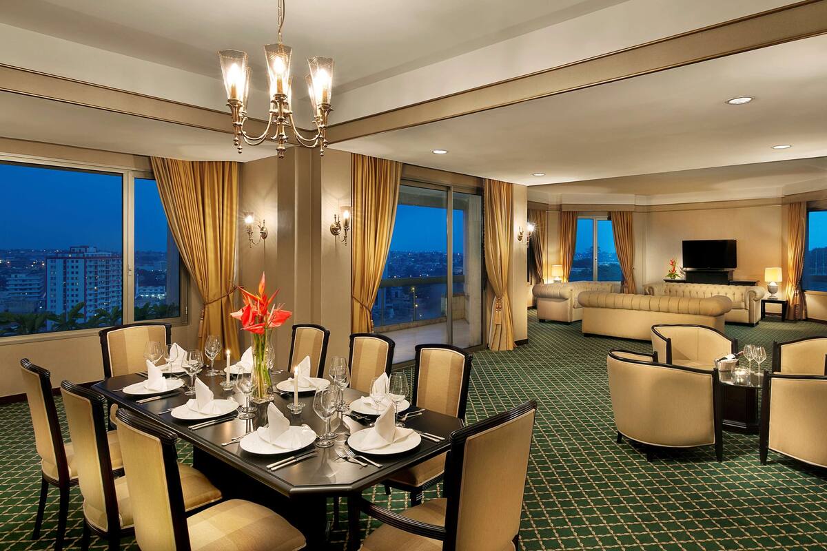 A hotel room with a dining area set for four, a living area with a sofa and armchairs, and a large window with a city view.