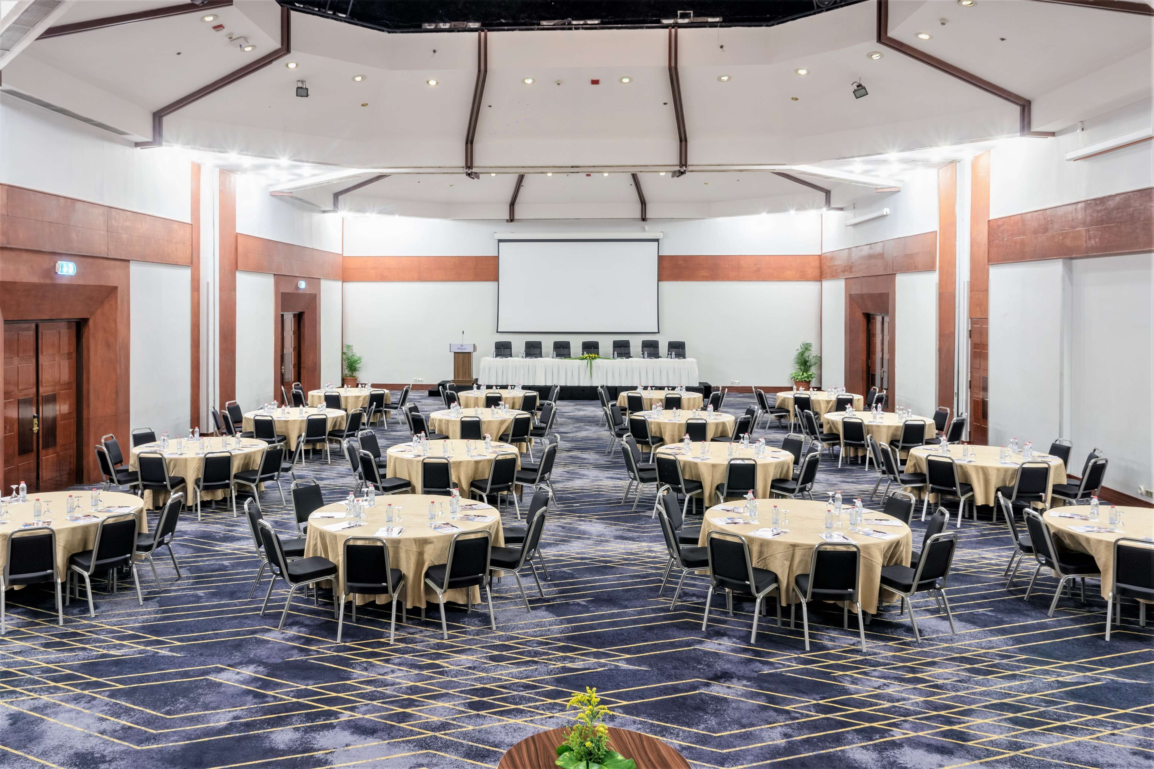 A conference room with multiple round tables, each with chairs, a stage with a projector screen, and a carpeted floor.