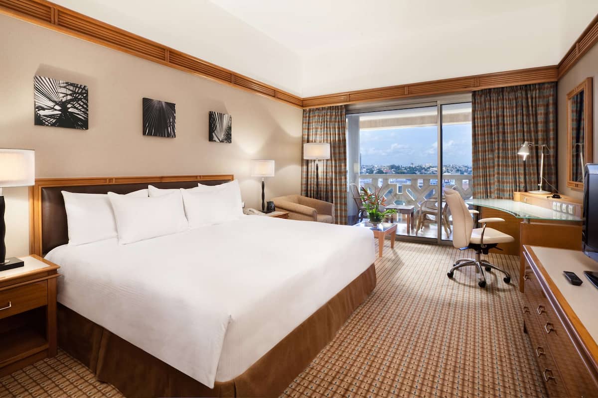 A hotel room with a large bed, a desk with a chair, a TV, and a balcony with curtains.