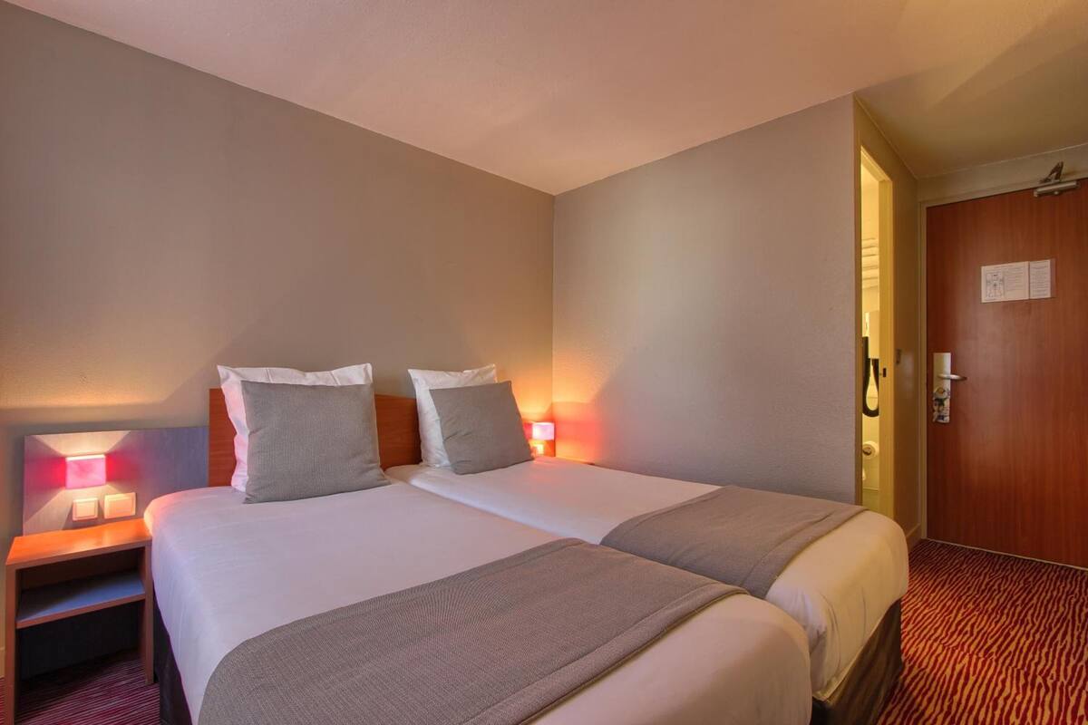 A hotel room with two beds, a nightstand, a lamp, a door, and a carpeted floor.