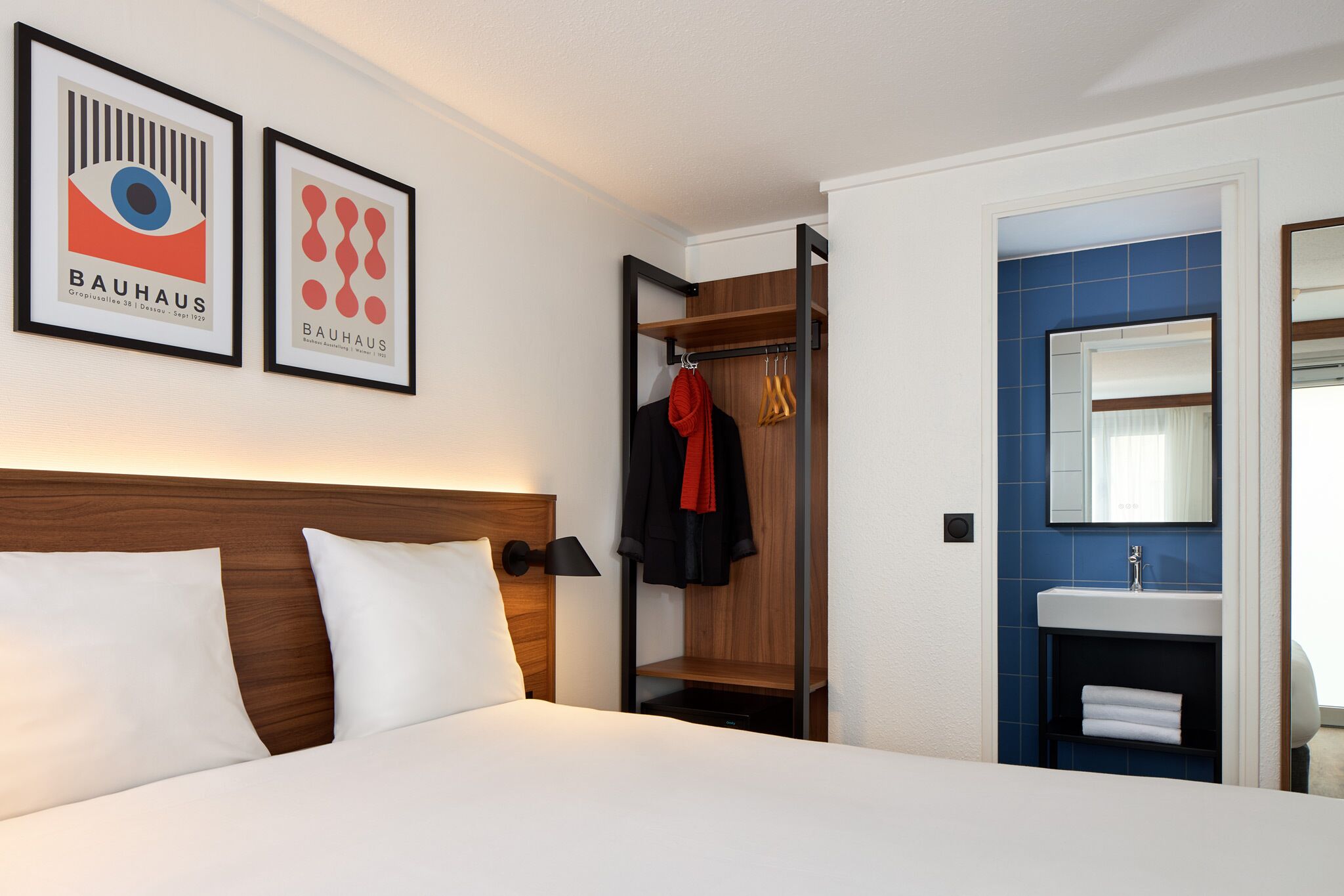 A hotel room with a bed, a nightstand, a wooden headboard, a wall-mounted shelf with clothes, a mirror, and a bathroom with a sink and blue tiles.