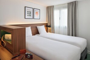 A hotel room with a wooden headboard, two beds, a desk with a lamp, a camera, and a handbag.