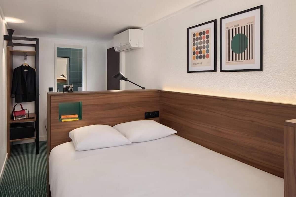 A hotel room with a wooden headboard, a bed with white linens, a bedside table, a lamp, a mirror, a wardrobe, and two framed artworks on the wall.