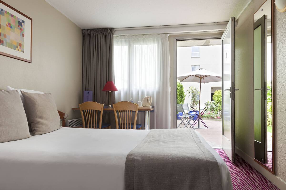 A hotel room with a bed, a table with chairs, a lamp, and a view of the outside.