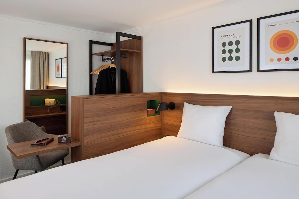 A hotel room with a wooden headboard, a bed with white linens, a desk with a chair, a hanging coat rack, and two framed pictures on the wall.