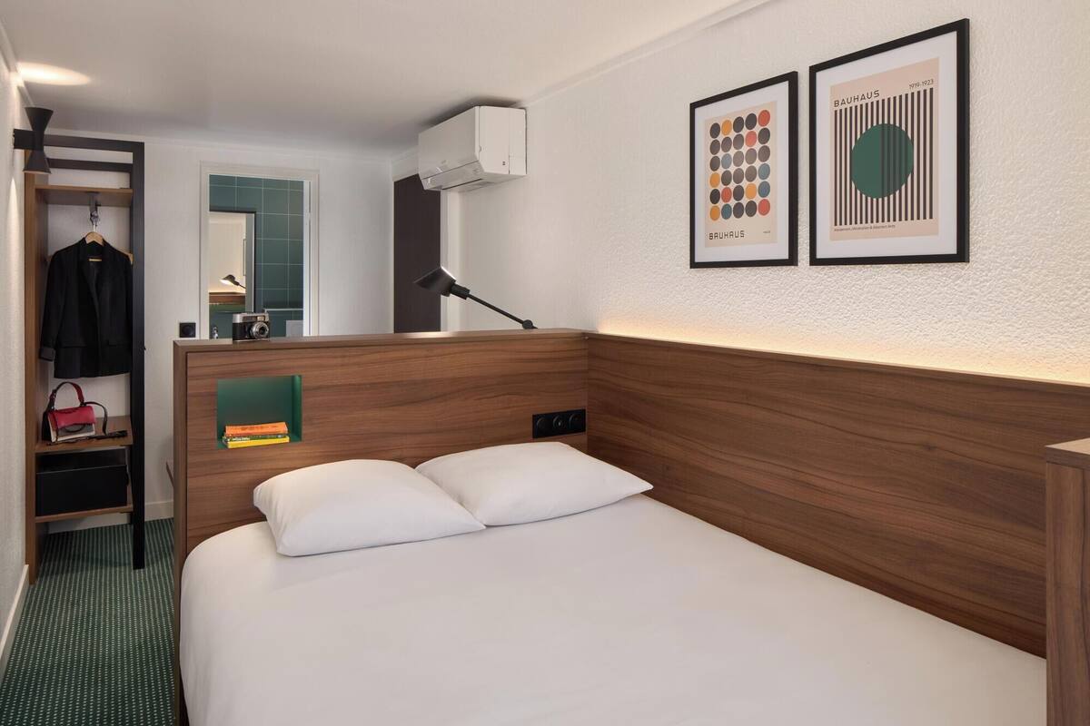 A hotel room with a wooden headboard, a bed with white linens, a bedside table, a lamp, a wardrobe, and two framed artworks on the wall.