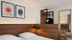 Quarto de hotel com cabeceira de madeira, cama com roupa de cama branca, luminária verde fixada na parede, um espelho e duas obras de arte emolduradas na parede.