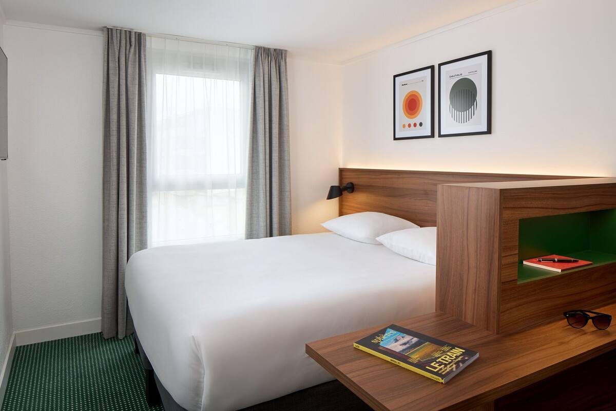 A hotel room with a neatly made bed, a bedside table with a book and glasses, and two framed artworks on the wall.