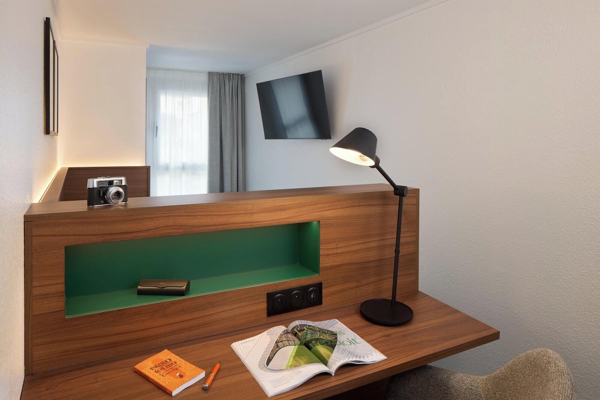 A hotel room with a wooden desk, a green storage compartment, a black desk lamp, a camera, and a magazine.