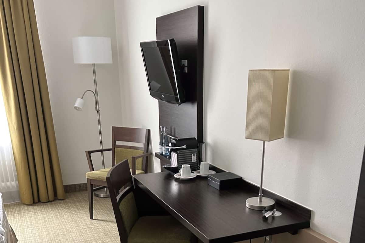 Une chambre d’hôtel avec un bureau en bois foncé, un téléviseur à écran plat fixé au mur, une chaise, une lampe et une machine à café.
